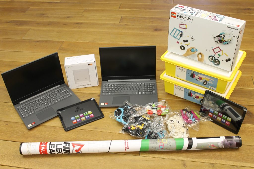 Na podłodze znajdują się dwa laptopy, dwa tablety, pudełko z routerem Wi-Fi, trzy pudełka z klockami Lego, przed nimi leży mata zwinięta w rulon.